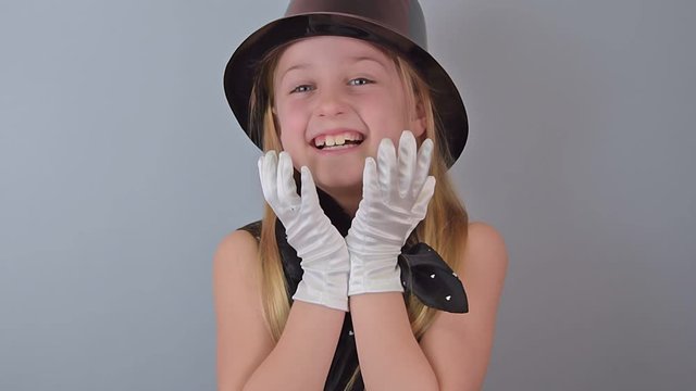 Funny positive little girl magician in a black hat and white gloves shows a terrifying laugh on a gray background. Concept of gloating and black humor