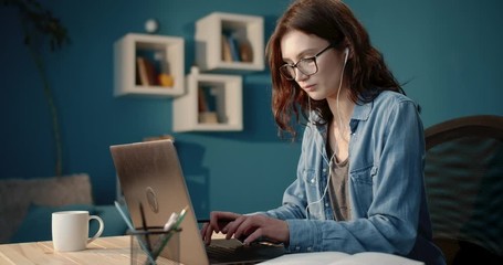 Attractive brunette student in eyeglasses and denim shirt using laptop for studying at home. Young lady with curly hair in headphones reading book and taking notes. - Powered by Adobe