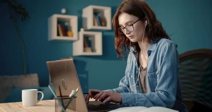 Attractive brunette student in eyeglasses and denim shirt using laptop for studying at home. Young lady with curly hair in headphones reading book and taking notes.