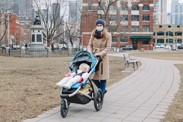 Young Caucasian mother in a surgical mask walking with baby outdoor in Toronto. Protective face mask precaution against new Chinese atypical pneumonia COVID-19 epidemic virus disease.
