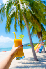 First-person view. Girl holds a glass cup of cold mango fresh on the background of a sandy tropical beach. White sand and palm trees. Fairytale vacation