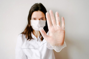 On a light background, a brunette woman in a white medical coat with gray accents and a protective mask on her face closeup in the center of the frame. Right hand in focus