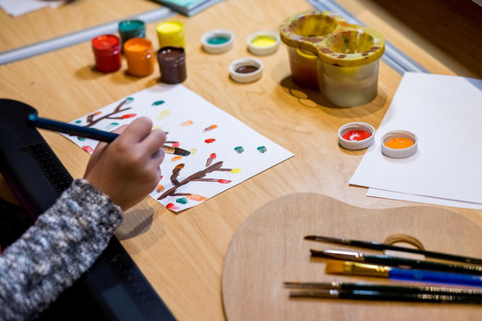 Kid hands painting at the table with gouache,art supplies, selective focus.Learning painting concept, paint brush and box with watercolors on wooden table with splashes,  workplace for children kids.