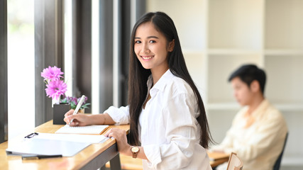 Fototapeta premium Photo of young beautiful woman working as accountant writing on document file while sitting at the wooden counter bar with colleague in office as background.