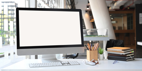 Photo of white blank screen computer monitor, wooden pencil holder, stack of books, wireless mouse and keyboard putting on white working table with modern office as background.