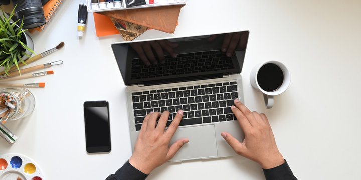 Top view image of graphic designer's hands typing on black blank screen computer laptop that putting on the white working table with coffee cup, black blank screen mobile and painting equipment.