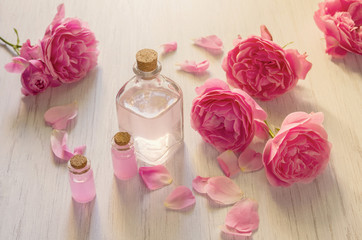 Rose water in glass bottles and pink rose flowers on wooden light background, aromatherapy concept.