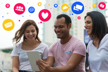 Smiling people sitting in park and looking at tablet with social media network icons above. Cheerful friends hanging out together. Communication and technology concept
