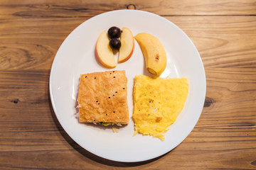 Top view of Breakfast menu: Omelette, Croissant sandwich, banana, apple and grapes in white plate on wooden table. Taipei hostel food service.