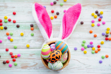 Top view of arrangement colorful easter eggs in a bowl and pink bunny ear,  decoration with candy and chocolate ornament on white wooden background. Happy Easter holiday background concept, copy space