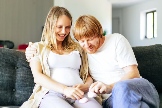 Happy Couple Expecting Daughter. Cheerful Future Mother And Father Playing With Pink Baby Shoes, Smiling, Laughing, Having Fun. Young Family Concept