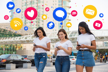 Joyful friends walking on street with smartphones and like, new message, comment media icons. Smiling young women holding modern phones during stroll. Technology concept