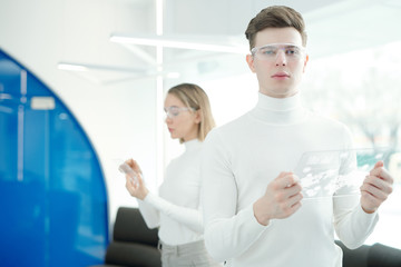 Portrait of serious handsome young man in modern goggles using futuristic tablet while analyzing...