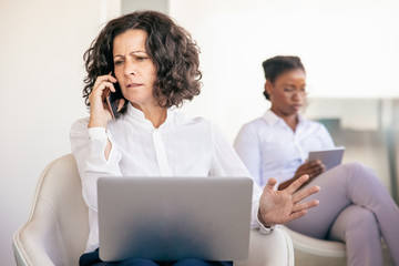 Annoyed businesswoman talking to partner on phone. Business woman sitting in armchair, using laptop and calling on cellphone in office lobby. Mobile negotiation concept