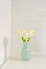 Close up vertical view of white calla lilies in green vase on table against painted brick wall