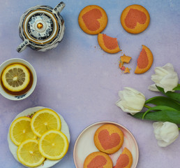 Flat lay a cup of tea , lemon slices, homemade cookies and white tulip flowers on a purple background. Top view