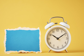 White vintage classic alarm clock with bells and empty blue torn paper sheet on the yellow...