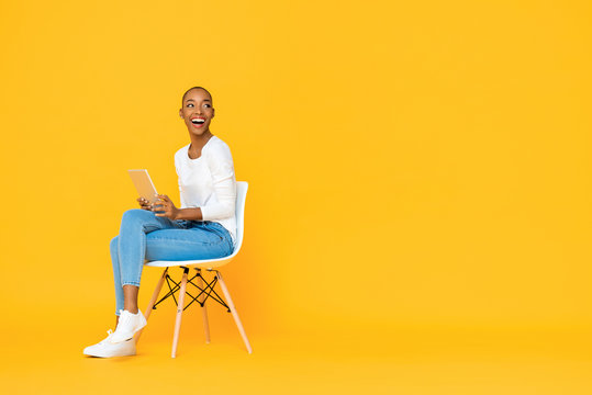 Trendy smiling African American woman sitting on a chair using tablet computer thinking and looking at empty space aside isolated yellow background