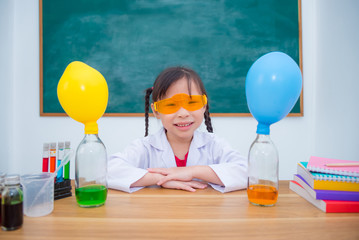 Little asian student wearing white coat sitting at dresk and smiles in front of blackboard at...
