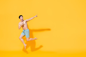 Fototapeta na wymiar Full length portrait of young happy shirtless Asian man in beach attire jumping in mid-air pointing two fingers at blank space beside in yellow isolated studio background