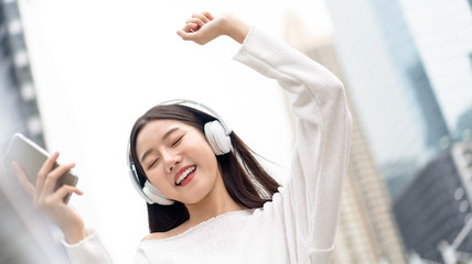 Happy smiling Asian girl wearing headphones listening and moving to the music against city building background