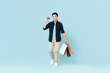 Handsome young Asian tourist man carrying bags shopping with credit card isolated on light blue...