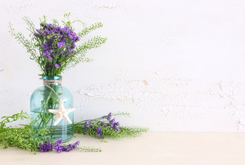 summer bouquet of purple field flowers in the nautical vase with seashell over wooden table and white old background