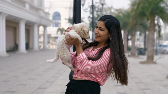 Beautiful young girl having fun with her cute little puppy in the city market in India. Attractive Indian female happily playing with her small dog while holding him gently in her arms - owner and ...