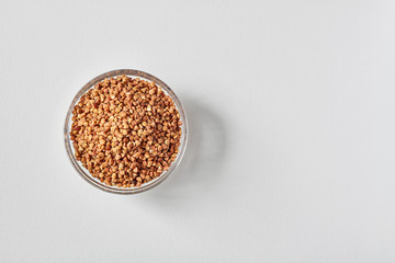Raw buckwheat groats in a transparent glass plate on white background. Closeup, copy space, top view