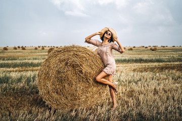 Naklejka na ściany i meble Beautiful young girl with long hair in sunnglasses and straw hat posing on a wheat field near hay bales. Happy brunette in summer dress