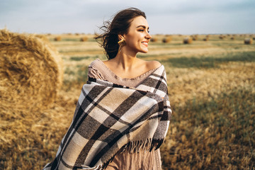 Portrait of a beautiful brunette in a dress and with a warm plaid. Woman enjoying a walk in a wheat field with hay bales