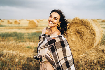 Portrait of a beautiful brunette in a dress and with a warm plaid. Woman enjoying a walk in a wheat field with hay bales © bedya