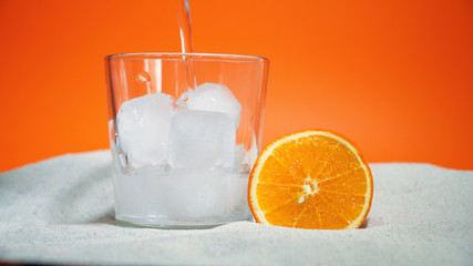 glass with ice and soda water and orange on sand on an orange background