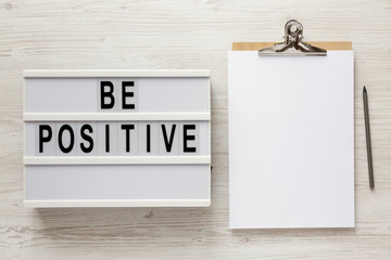 'Be positive' words on a lightbox, clipboard with blank sheet of paper on a white wooden background, top view. Overhead, from above, flat lay.