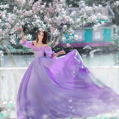 Fototapeta premium Portrait of amazing cute young woman with dark brown long hair in long purple dress with large flyingskirts stands under blooming white magnolia flowers against background of lake and blue fairy house