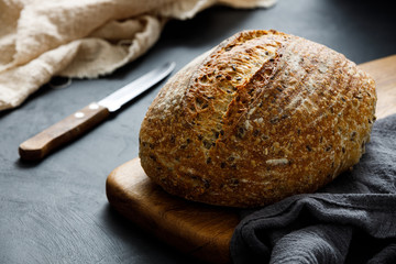 Round loaf of freshly baked sourdough bread with knife on cutting board. Artisan bread with seeds on dark table. Rustic sourdough bread.