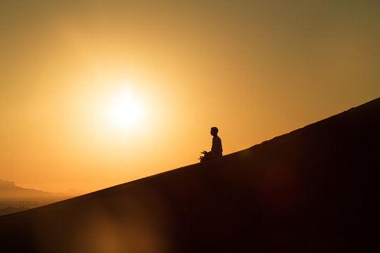 Silhoutte conceptual shot of young man meditating in the desert during sunrise on a sand dune; peaceful meditation in isolated place 