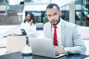 Serious confident business consultant waiting customer in office cafe. Young handsome man in office suit sitting at table in cafe, using laptop and staring into distance. Workspace concept
