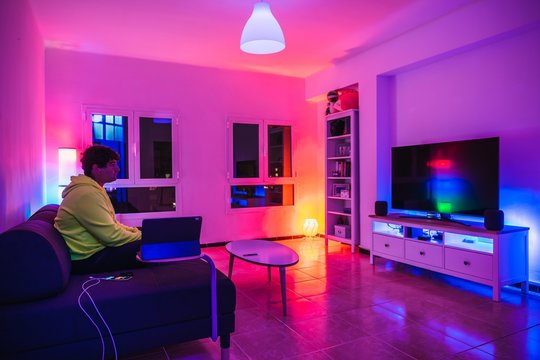 Young spanish man sits on it’s couch while sitting in front of tv and with his smart home system with all the lights on with different colors