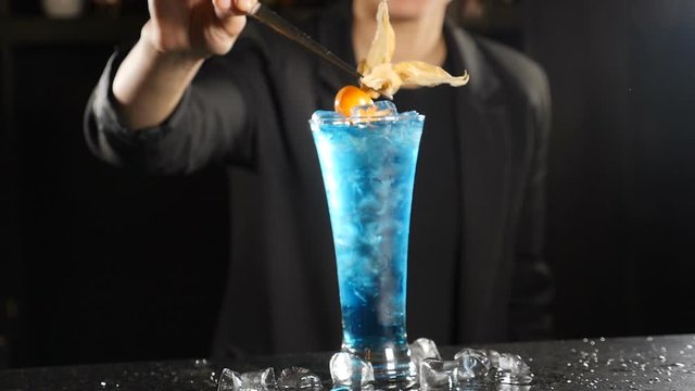 Young female bartender decorating bright blue cocktail with winter cherry in slow motion. Bar in nightclub. Cocktail party. Full hd