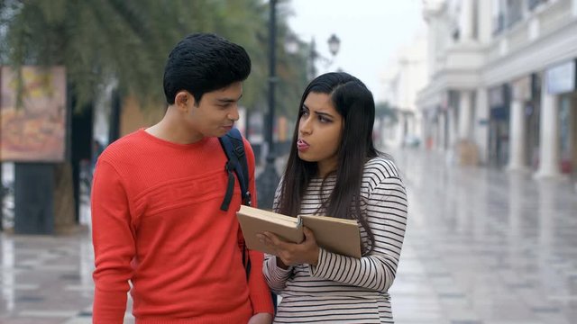 Cute Indian girl enjoying reading a book with her boyfriend - lifestyle concept. A young attractive couple happily blowing bubblegum candy bubble while reading a book outside a cafeteria - leisure ...