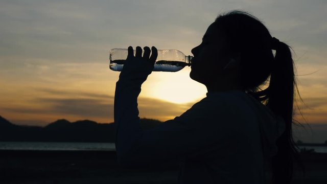 Silhouette athlete woman runner drinking water after running fitness workout at the beach in the last sunlight. fitness and healthy lifestyle concept. Slow Motion Shot.