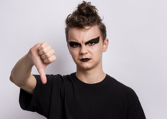 Teen boy with spooking make-up giving thumb down gesture looking with negative expression and disapproval. Teenager in style of punk goth dressed in black doing bad signal. Child on grey background. - 329757415