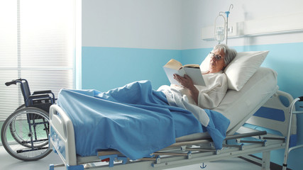 Senior woman lying in bed at the hospital and reading - obrazy, fototapety, plakaty