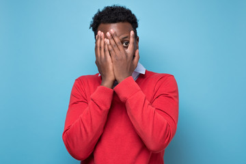 Young african american man in red sweater isolated on blue studio background peeping through fingers, hiding cover face with hands, looking terrified