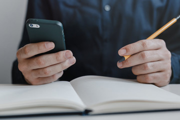 Closeup of the man's hands holding mobile phone and a pencil between fingers. Notebook on the table. Young businessman working in an office. Business, finance, industry and job concept.