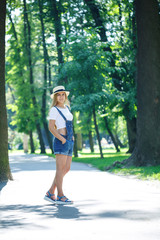 young beautiful girl in denim overalls and a light hat walking in the park