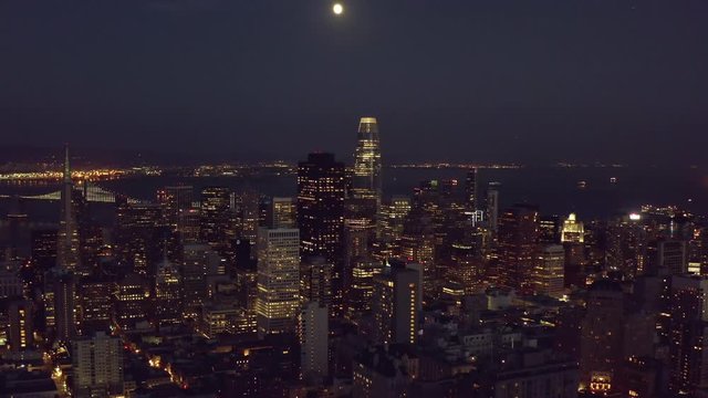 Aerial, tilt up, drone shot overlooking downtown San Francisco cityscape, revealing the full moon, during night time, in California, United States