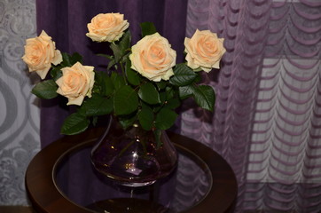 Against the background of purple textile, on a round glass table in a beautiful vase there is a bouquet of beautiful fresh fragrant orange roses with green leaves.