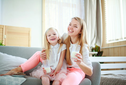 Young Mother With Child Daughter At Home On Couch Drinking Milk And Cookies, Laughing And Talking Together